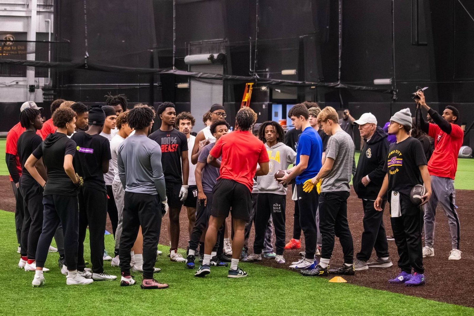 A BSP coach in a red shirt instructs a large group of athletes on an indoor turf field